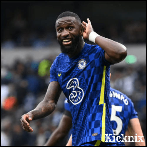 Antonio Rüdiger in a Chelsea jersey celebrating with his hand cupped to his ear, gesturing 'I can't hear you' in response to the crowd.